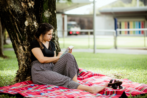 A young person sitting on a picnic blanket under a tree looking at the back of a book